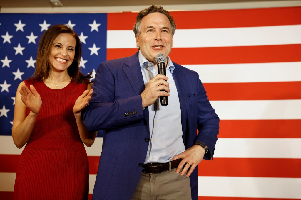 Dave McCormick speaking in mic with wife Dina Powell-McCormick at his side with American flag behind.