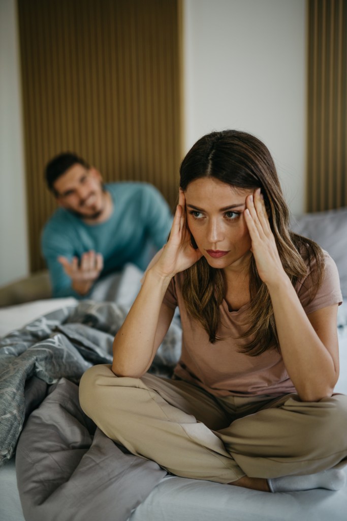 Young couple having marital problems in the bedroom, with upset woman on the bed and arguing husband in the background