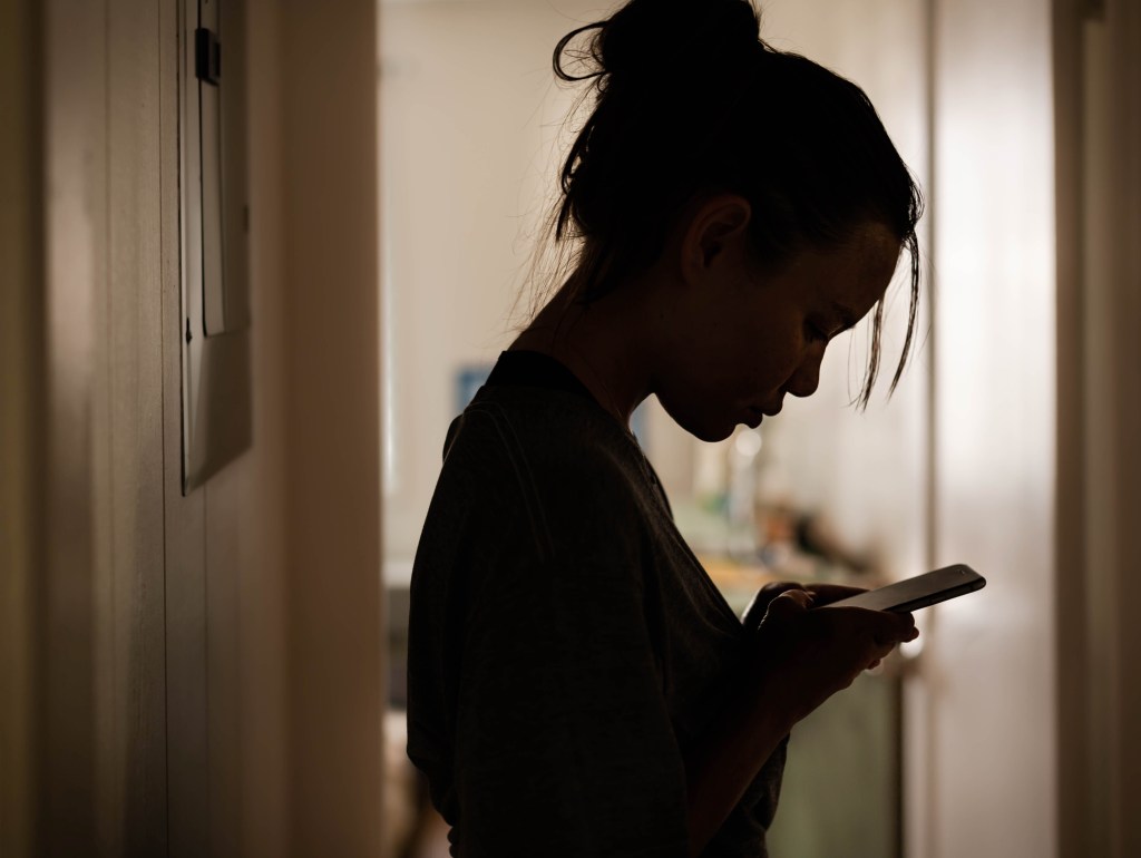 Young women, in shadow, looking at a phone in her hands. A door opens to a room behind her