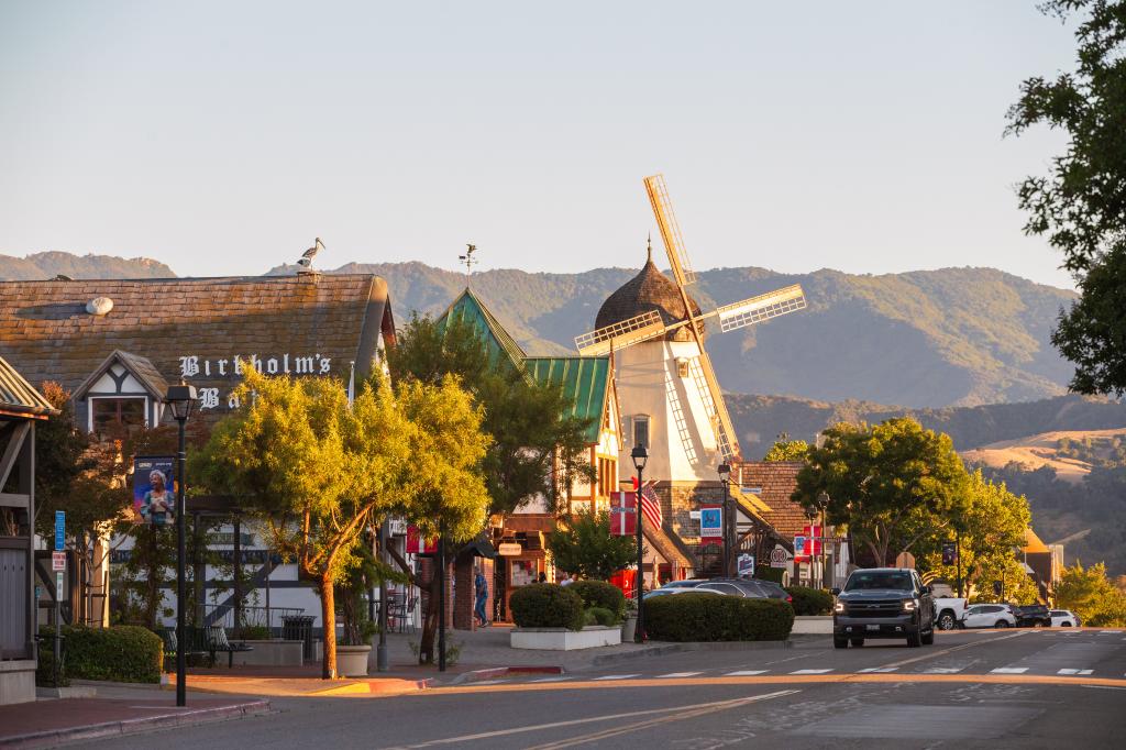 A street view of downtown Solvang with buildings and a windmill, surrounded by green spaces and mountains.