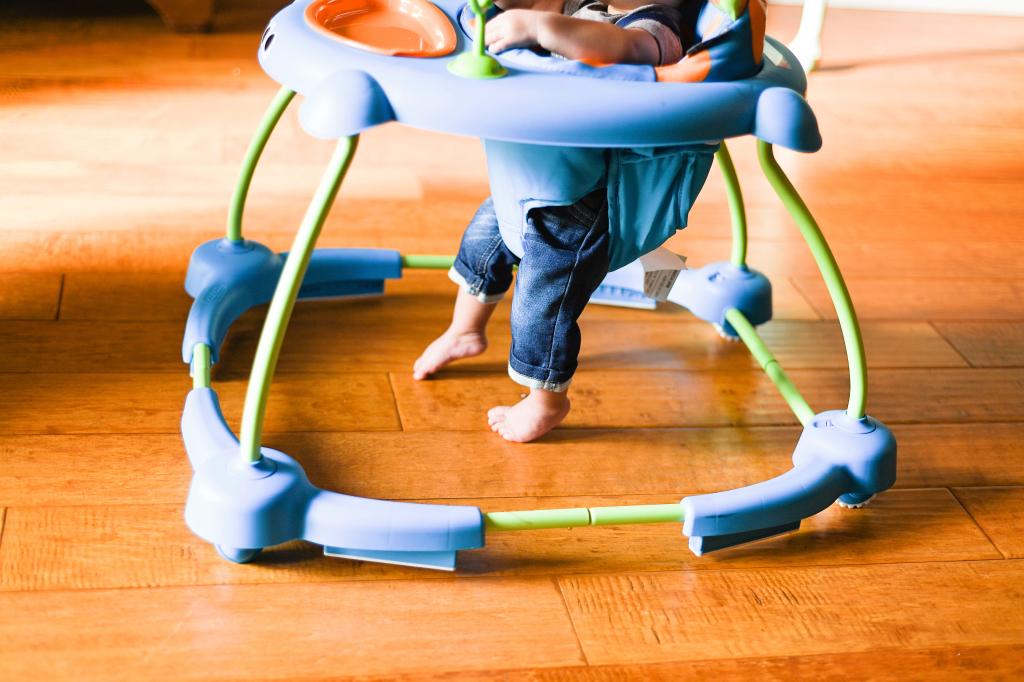 Baby's legs in a blue and green walker on a wooden floor, compliant with federal safety features