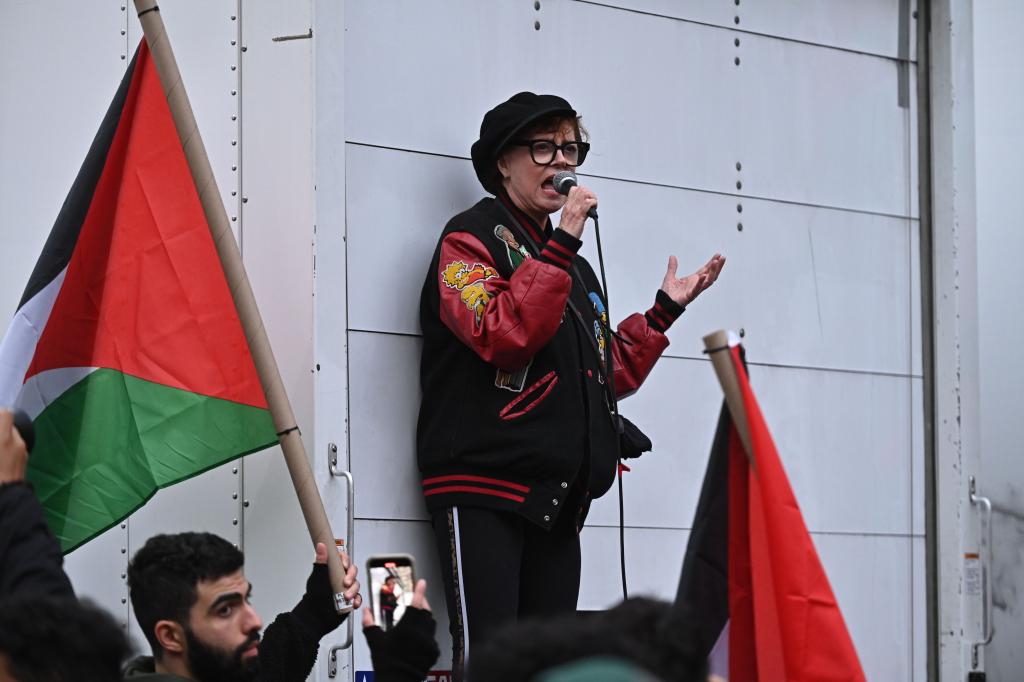 Susan Sarandon speaking into a microphone a a rally, next to a Palestinian flag