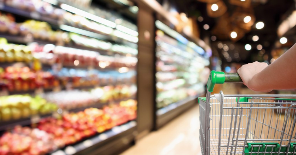 Woman's hand holding a supermarket shopping cart, with blurred background of fresh fruits and vegetables on grocery store shelves