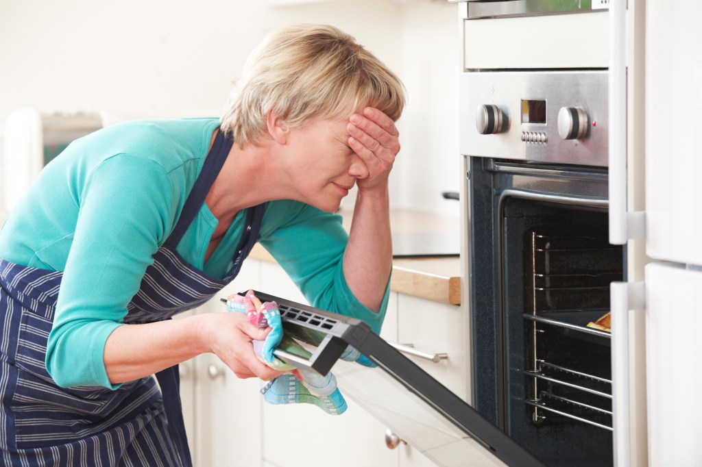 Woman covering her eyes while looking at disastrous meal in oven