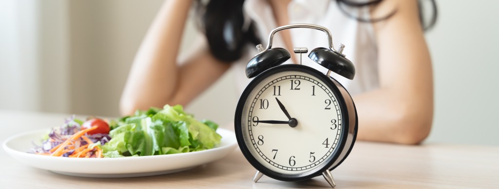 Intermittent fasting — which involves limiting eating to a certain daily window — is not for everyone. Researchers have identified pros and cons to this popular meal schedule. Here, a woman waits to eat next to a ticking alarm clock.