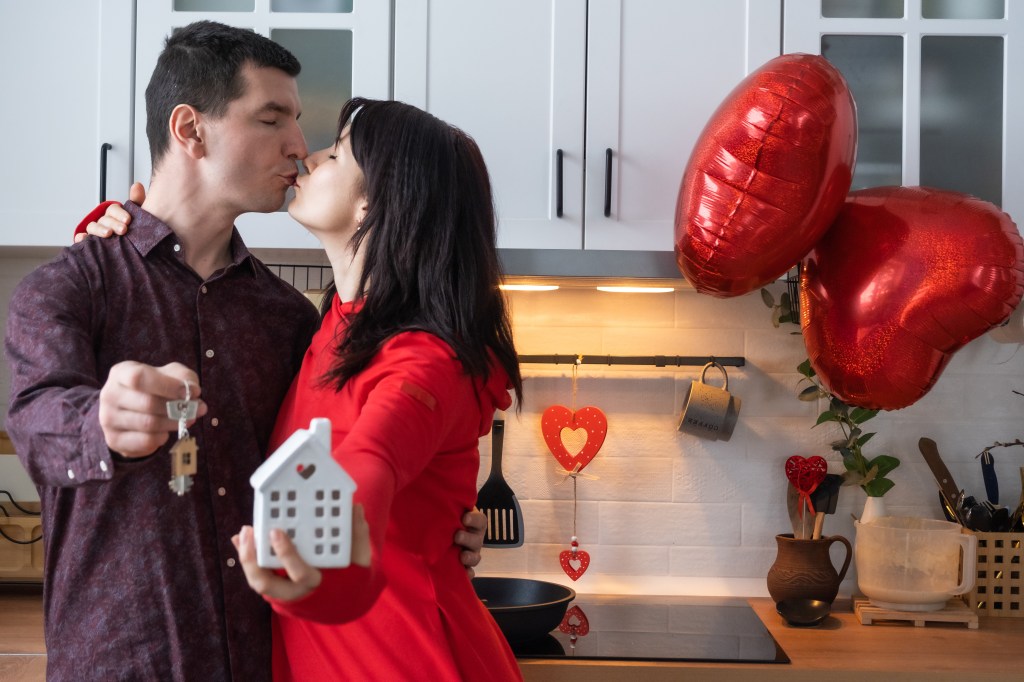 A man and woman kissing in a kitchen, holding house keys and red heart balloons