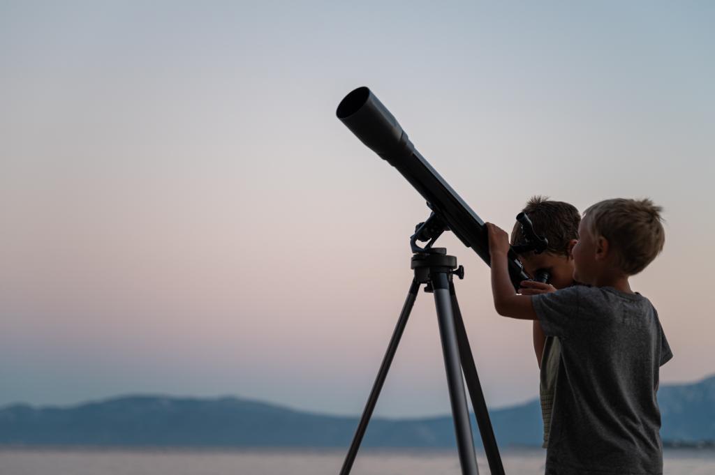 Two toddler brothers looking at the stars using a telescope by the sea on a summer night