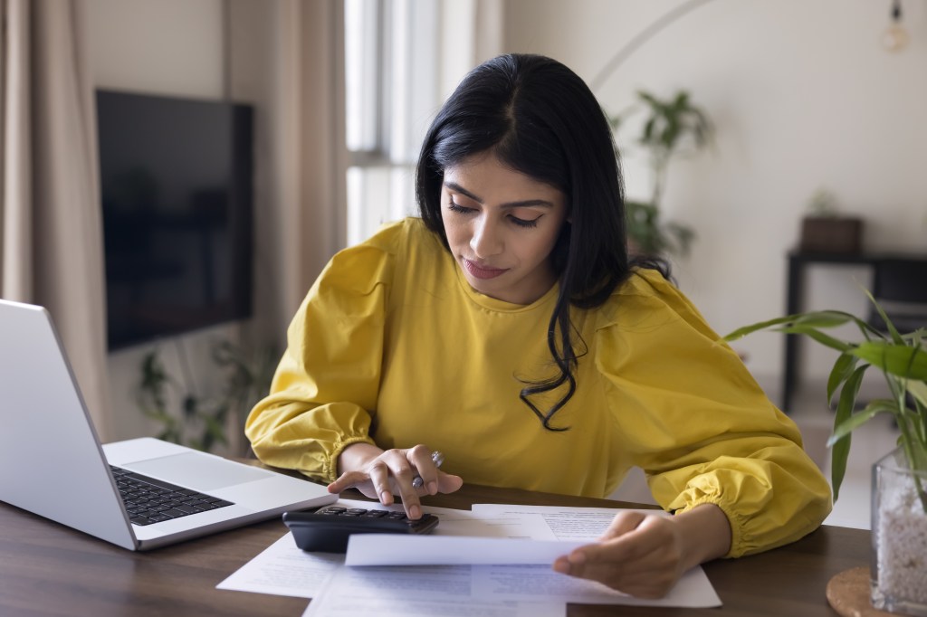 accountant woman checking financial documents, counting budget, income, expenses on calculator, sitting at home workplace table with laptop. 