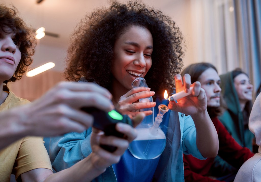 girl lighting marijuana in the glass bong, relaxing with friends on the sofa at home. Young people playing video games and smoking weed. 