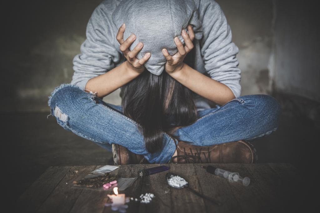 Woman sitting on the floor with a syringe, powdered drugs, pills and other supplements seen in front of her, illustrating the concept of drug addiction. She is seen with hood over head and head in hands.