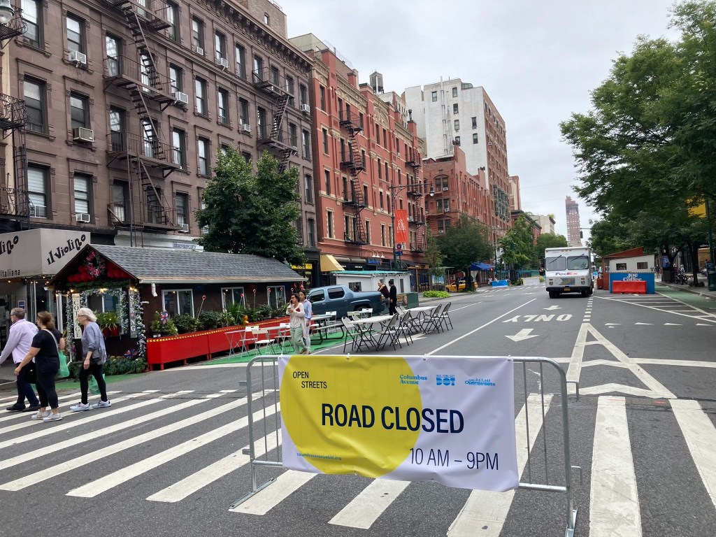 columbus ave from 68th to 77th closed to traffic with a medal gate and a yellow and white sign readingroad closed there is a dinning shed with several gtables and chairs in the empty roadway. further in the background there is a fed ex truck 