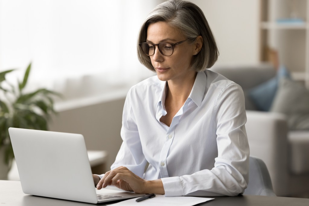 Focused pretty mature business woman in elegant glasses typing on laptop, working at computer in home office, using Internet technology for freelance job communication