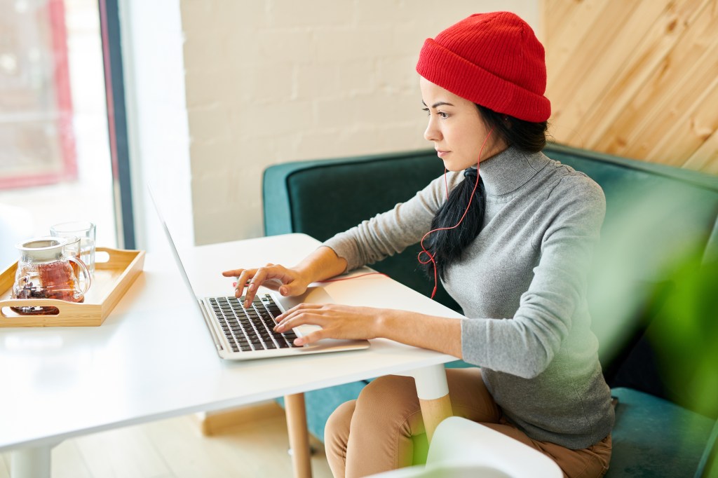High angle portrait of modern young woman wearing beanie hat using laptop doing freelance work in cafe and listening to music