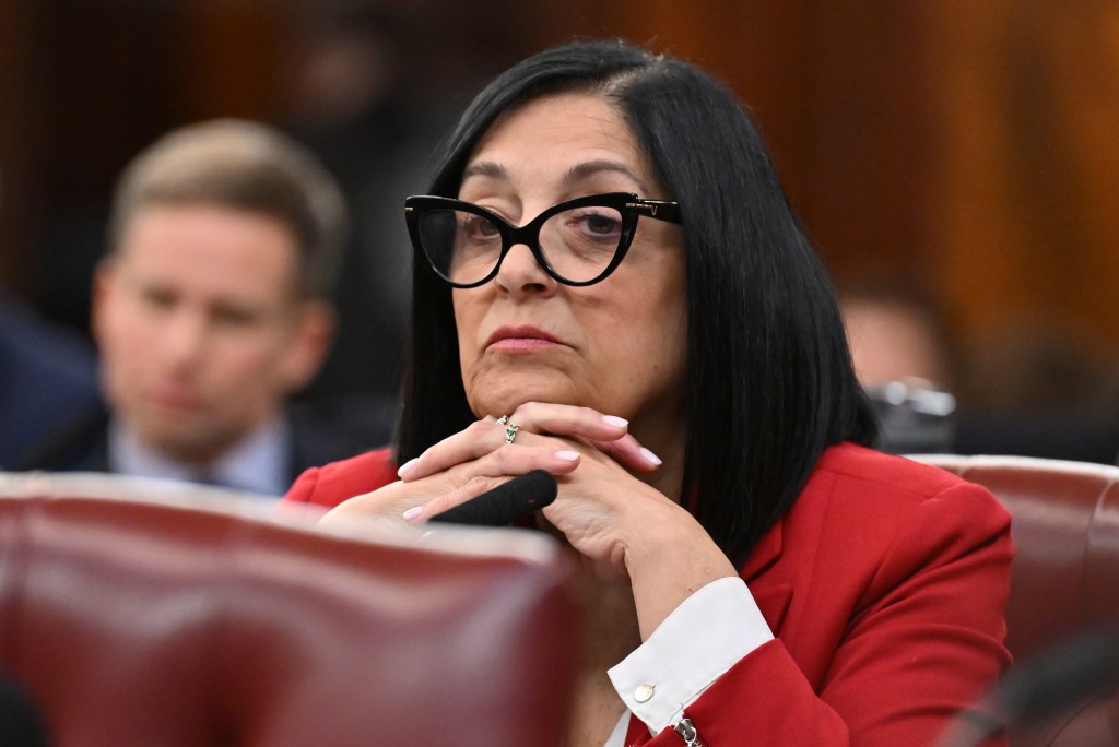 Joann Ariola in a red jacket and white shirt with black rimmed almost cats eye glasses sitting with her hands intertwined under her chin