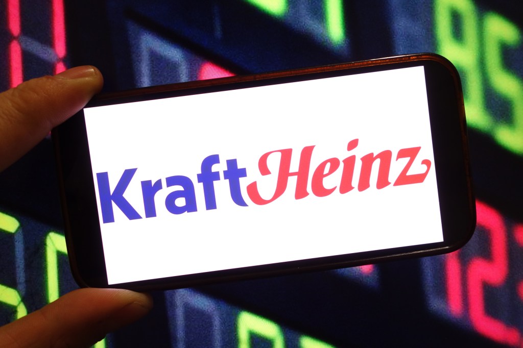 A hand holding a smartphone displaying the Kraft Heinz company logo, symbolizing their projected release of additional Crystal Light Vodka Refresher flavors in 2026.