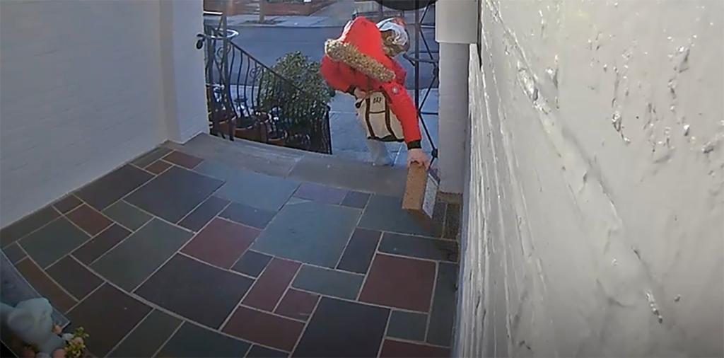 Woman  in an orange parka with the hood up, carrying two bags reaches out to grab a box from the multicolored slate front porch.