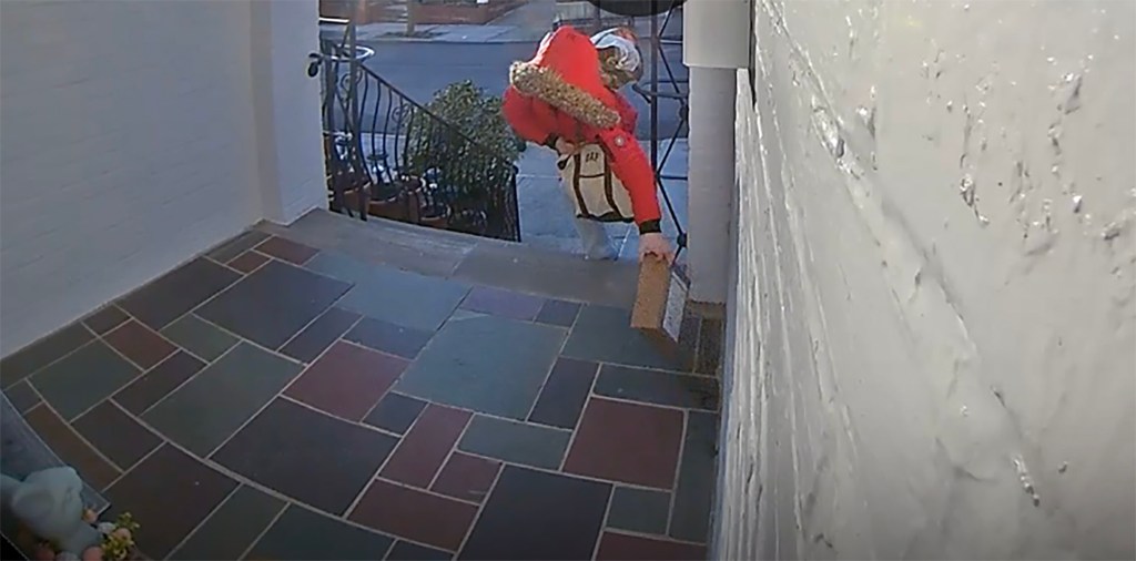 Woman  in an orange parka with the hood up, carrying two bags reaches out to grab a box from the multicolored slate front porch.