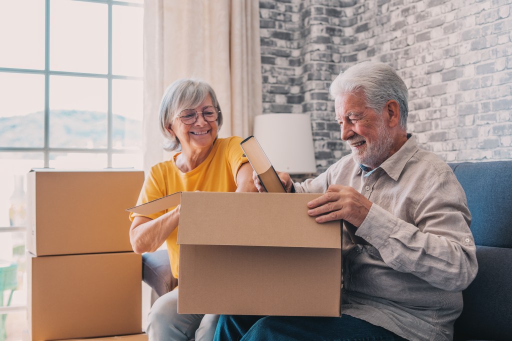 Relaxed mature married couple in love resting on couch among paper cardboard boxes, taking break, pause, hugging, talking, enjoying being in new home. Real estate concept