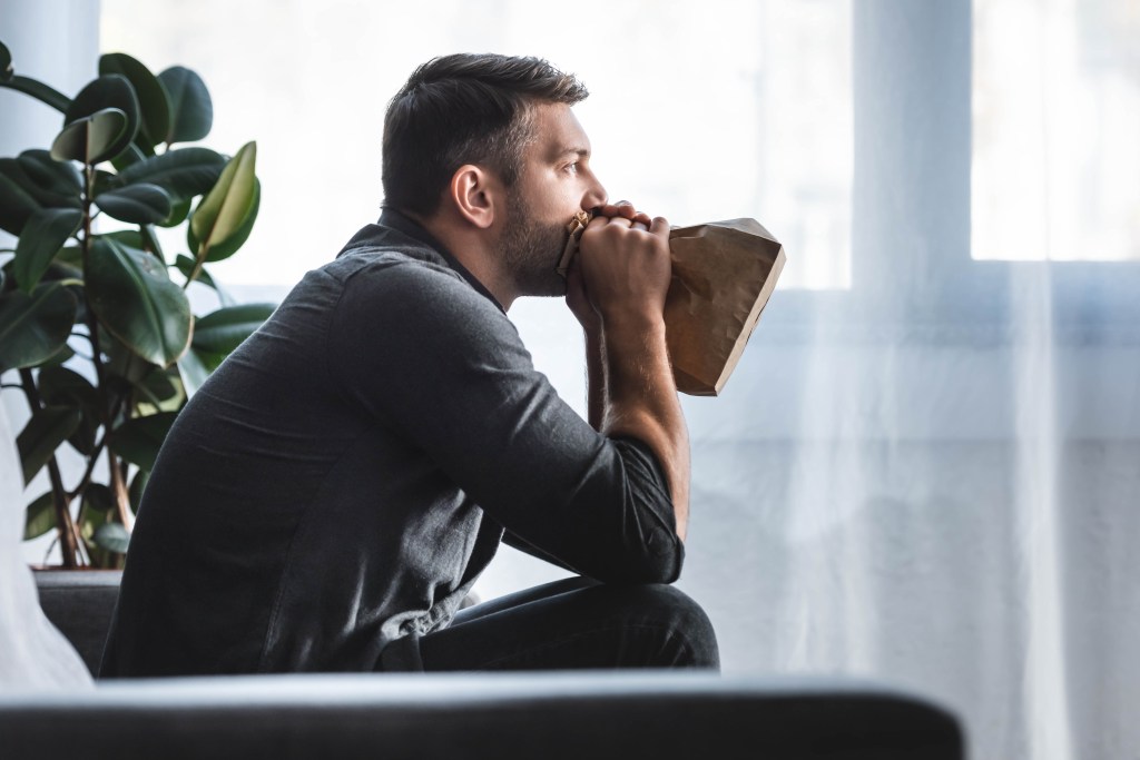 Side view of a worried man experiencing a panic attack, breathing into a paper bag in his apartment