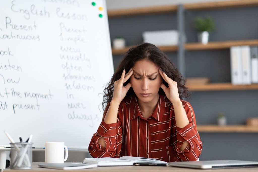 Exhausted young teacher touching her forehead and temples, suffering from a headache at her desk near a whiteboard