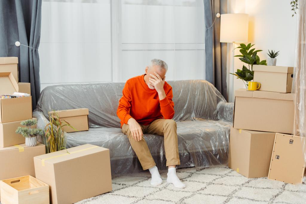 Upset senior man sitting on sofa feeling exhausted in new home living room with cardboard boxes packed with belongings on moving day