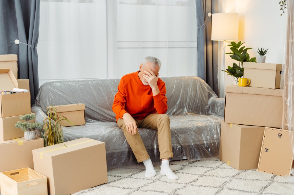 Upset senior man sitting on sofa feeling exhausted in new home living room with cardboard boxes packed with belongings on moving day