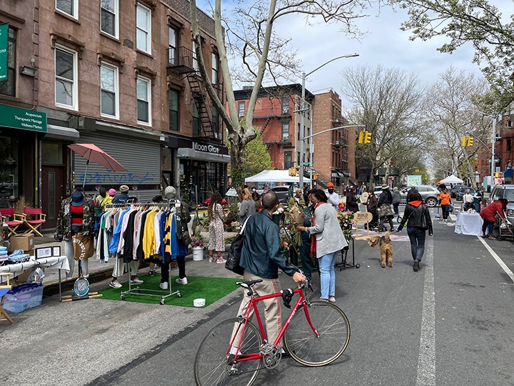 There are about 200 Open Streets operating through the DOT.  image is of people and dogs shopping and milling about on one of the closed streetw with a clothing vendor and a man walking a red bike
