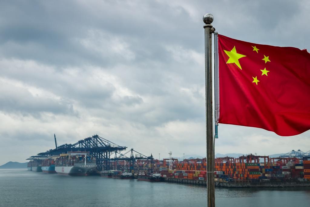 Chinese national flag in front of container ships, cranes, and stacked shipping containers at Yantian International Container Terminal, Shenzhen, China.