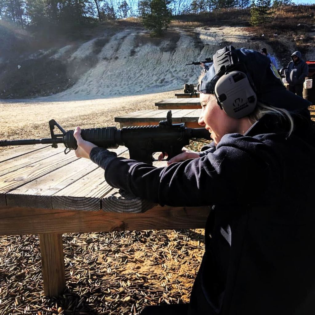 Reynolds at the firing range, shooting a rifle and wearing a black jacket and ear protection leaning on a wooden table.