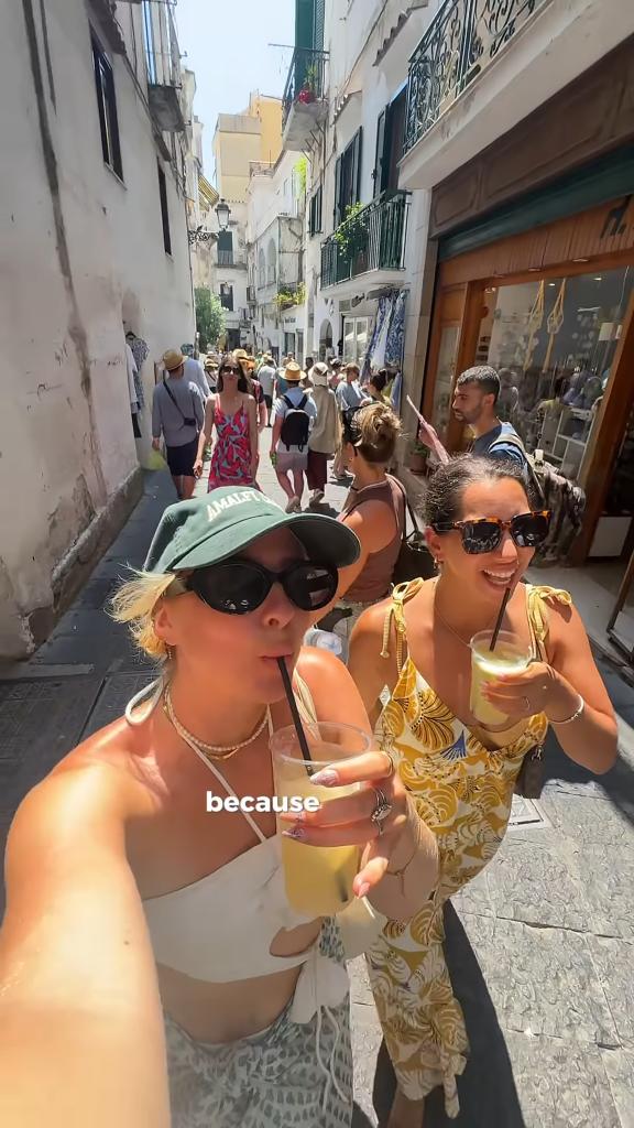A content creator and influencer among a group of tourists on a crowded street in a charming Italian town on a sunny day while drinking