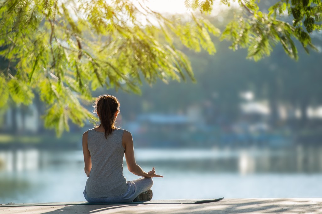 Beautiful woman sitting in a lotus position practicing yoga by the waterfront in a park