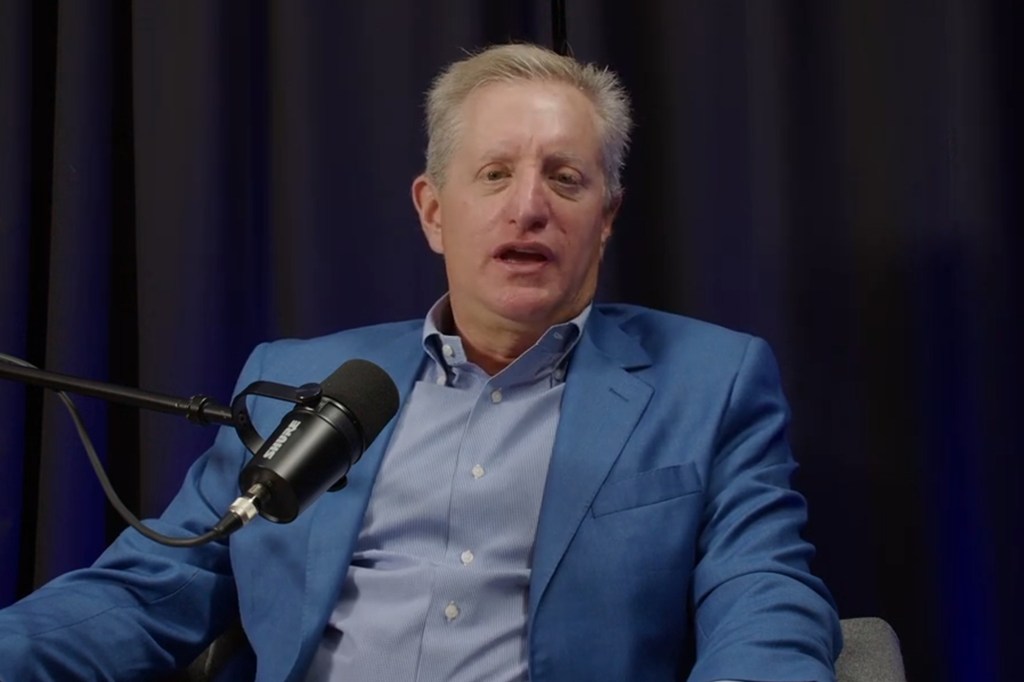 Steve Eisman, the 'Big Short Hero Investor', sitting in a chair while wearing a blue suit and holding a microphone during 'The Eisman Playbook' show