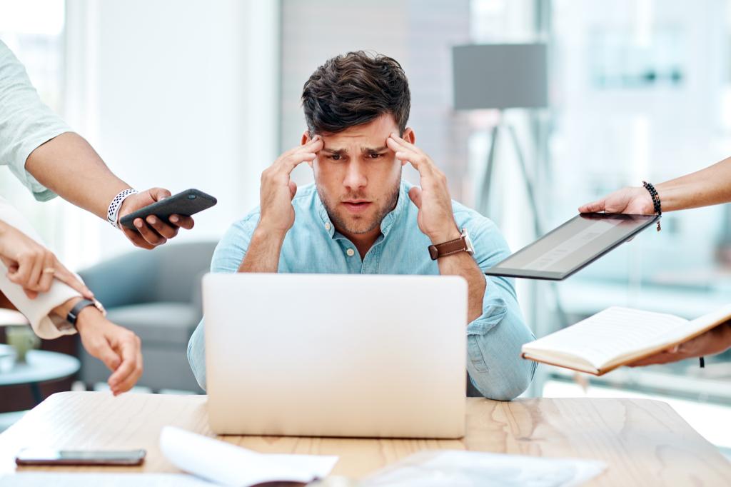 Stressed businessman multitasking at office desk with laptop, appearing overwhelmed and frustrated due to workload and time management issues