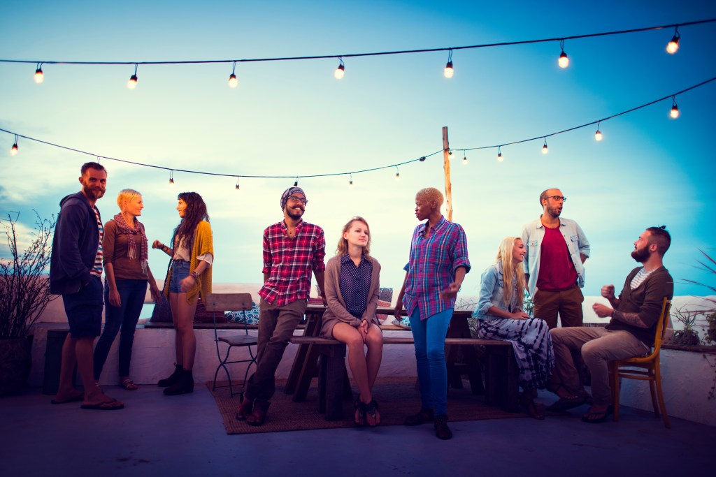 A group of diverse people, including celebrities Guillaume Samica, Alexander Gemignani, Judy Murray, Hana Hayes, standing around a table on a rooftop at sundown, near the beach