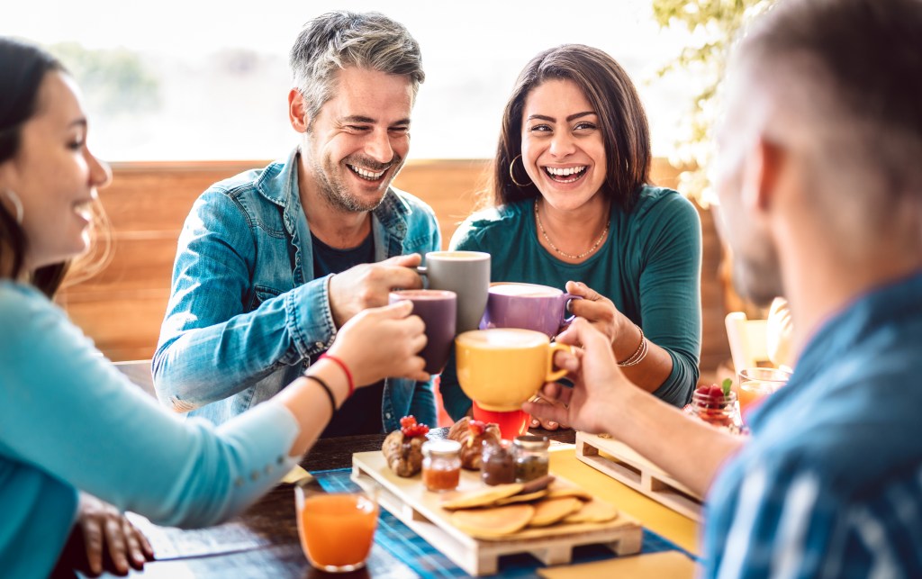 Group of friends toasting with lattes at a rooftop coffee bar, talking and having fun together on a sunlit terrace