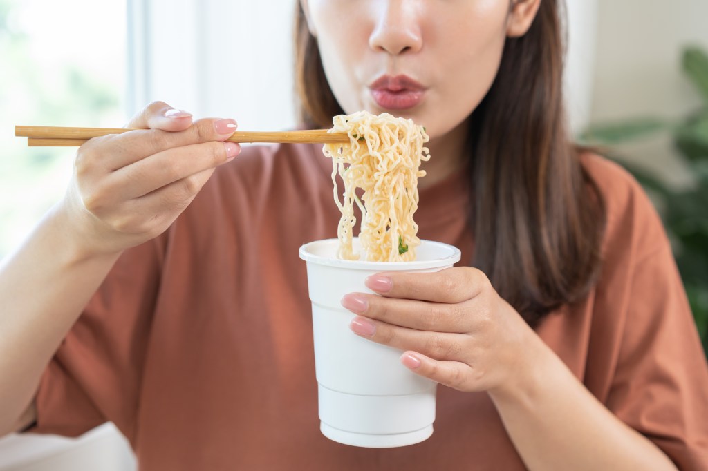 young student woman using chopsticks eating instant ramen, noodles soup from a cup, cooking meal fast food