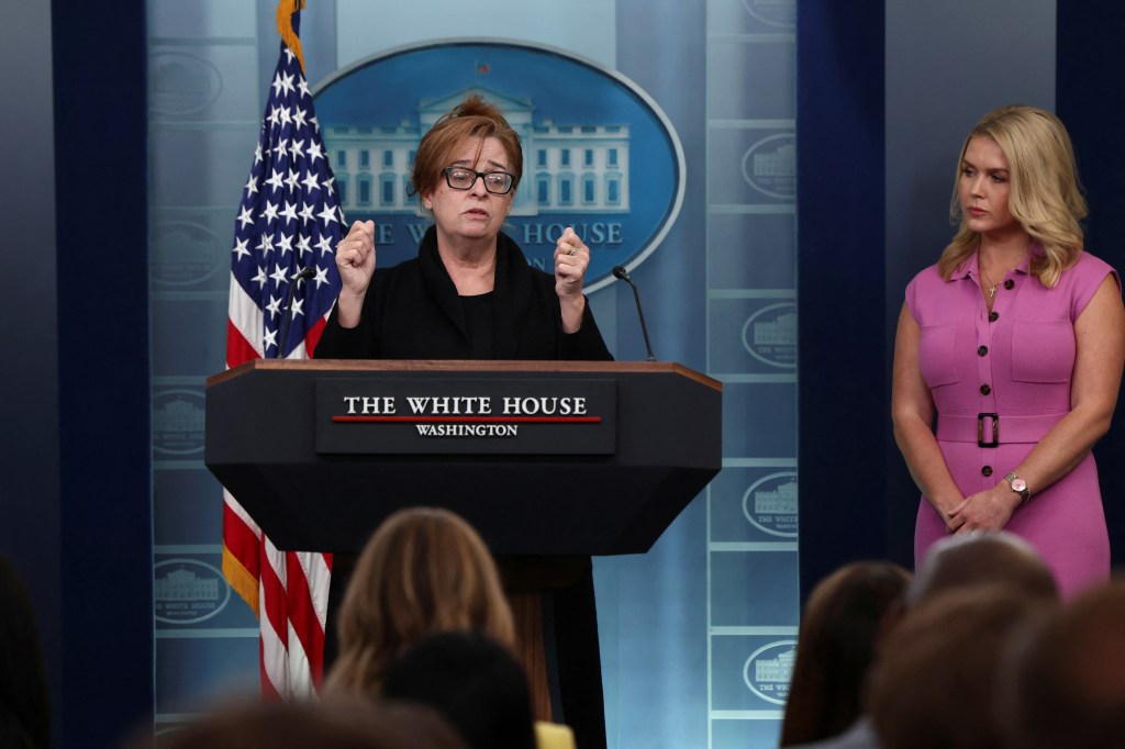 Patty Morin speaking at a press briefing at the White House with Press Secretary Karoline Leavitt standing by her side, American flag in the background.