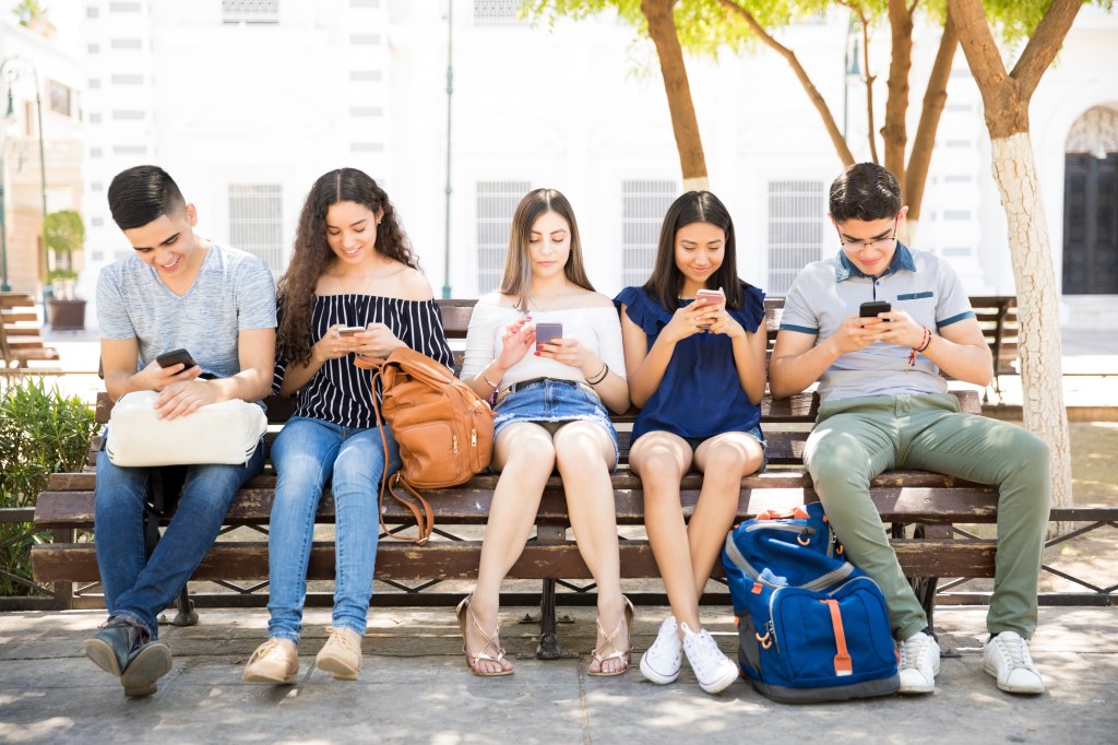 Portrait of teenage boys and girls with backpacks sitting outdoors on bench using their mobile phones