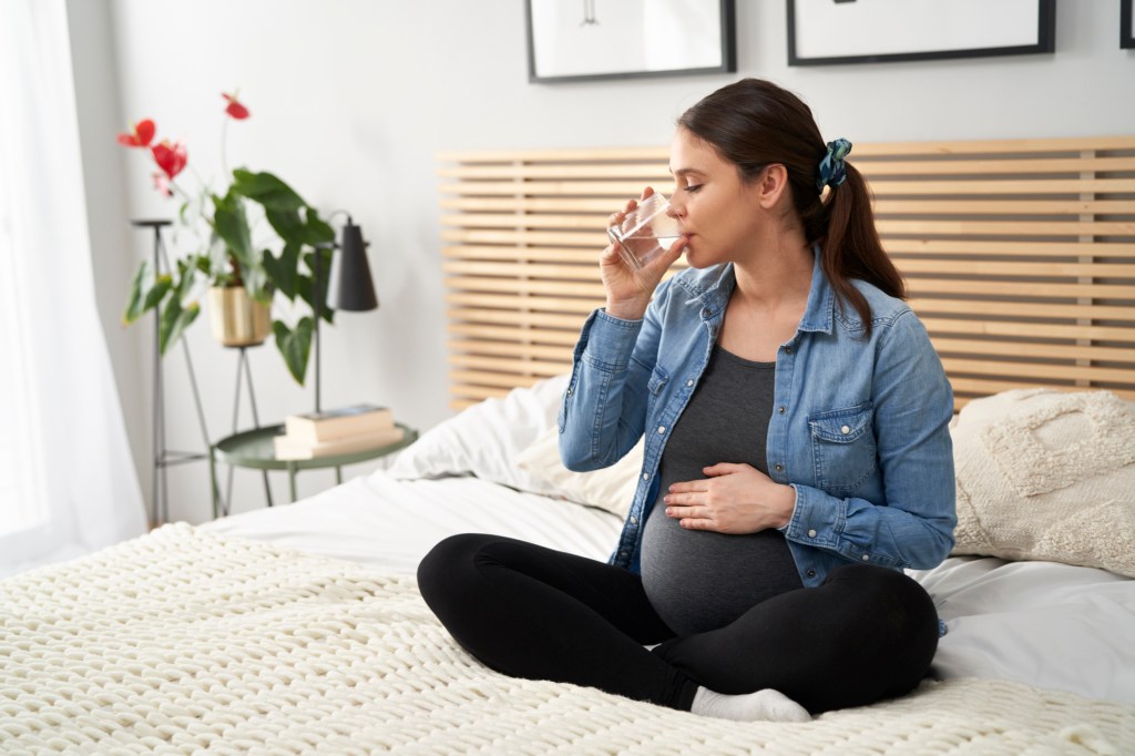 Ophélie Meunier, pregnant, sitting on a bed at home, drinking water