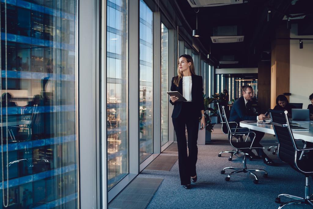 Confident female administrative manager in formal suit walking in office with digital tablet, looking at window