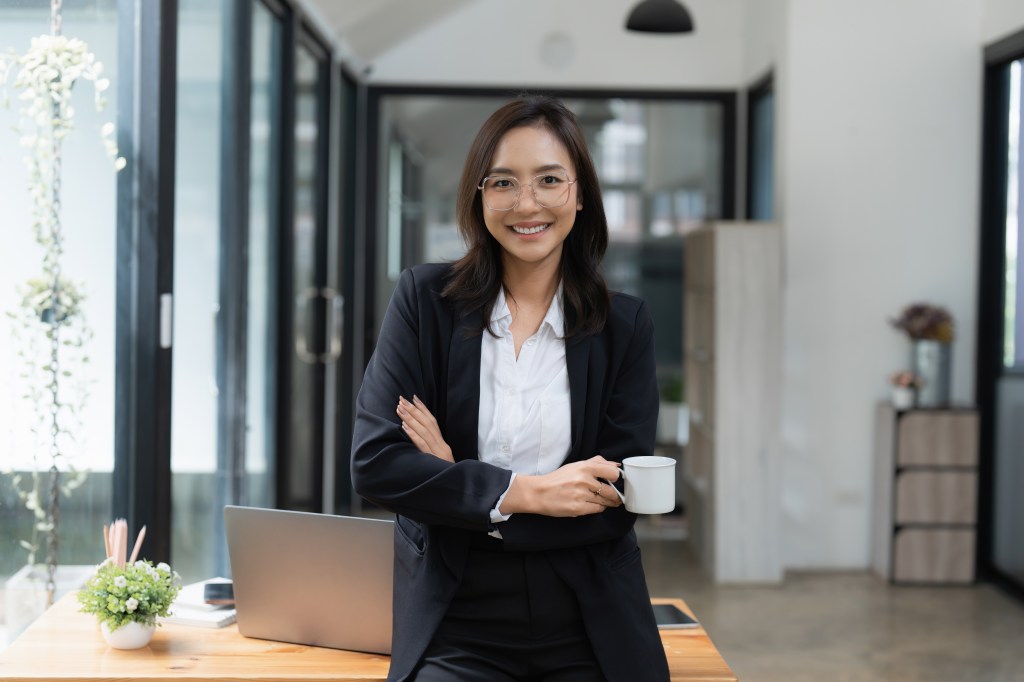 Smart Asian business woman in a suit, smiling and holding a cup in an office space