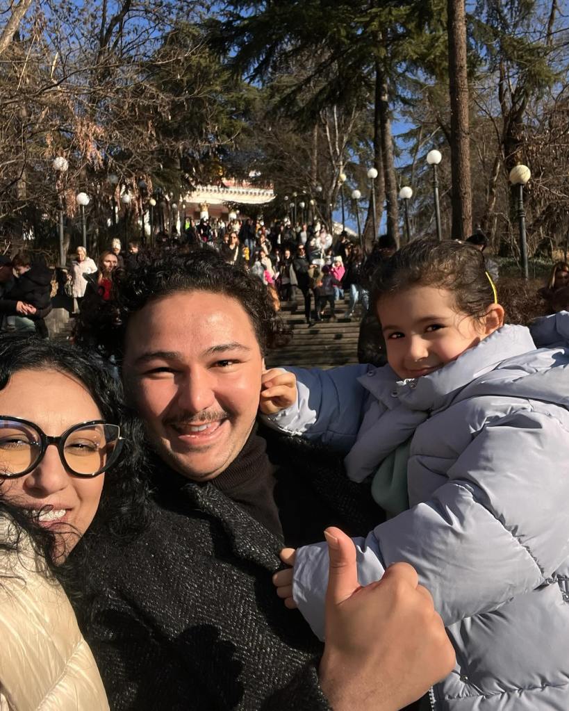 Rachvelishvili in dark rimmed glasses and a selfie giving a thumbs up with her husband, in a dark coat, and young daughter, in a light grey puffer jacket