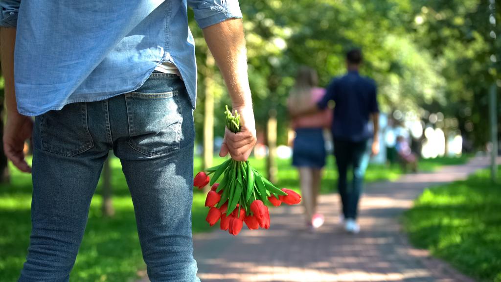 Man holding flowers looking disappointed as his girlfriend hugs another man