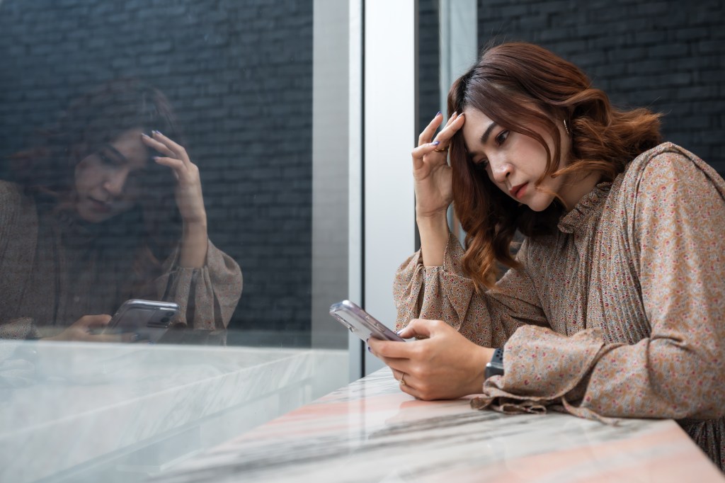 Depressed young woman looking at her smartphone and having a headache, feeling sad and worried about a problem