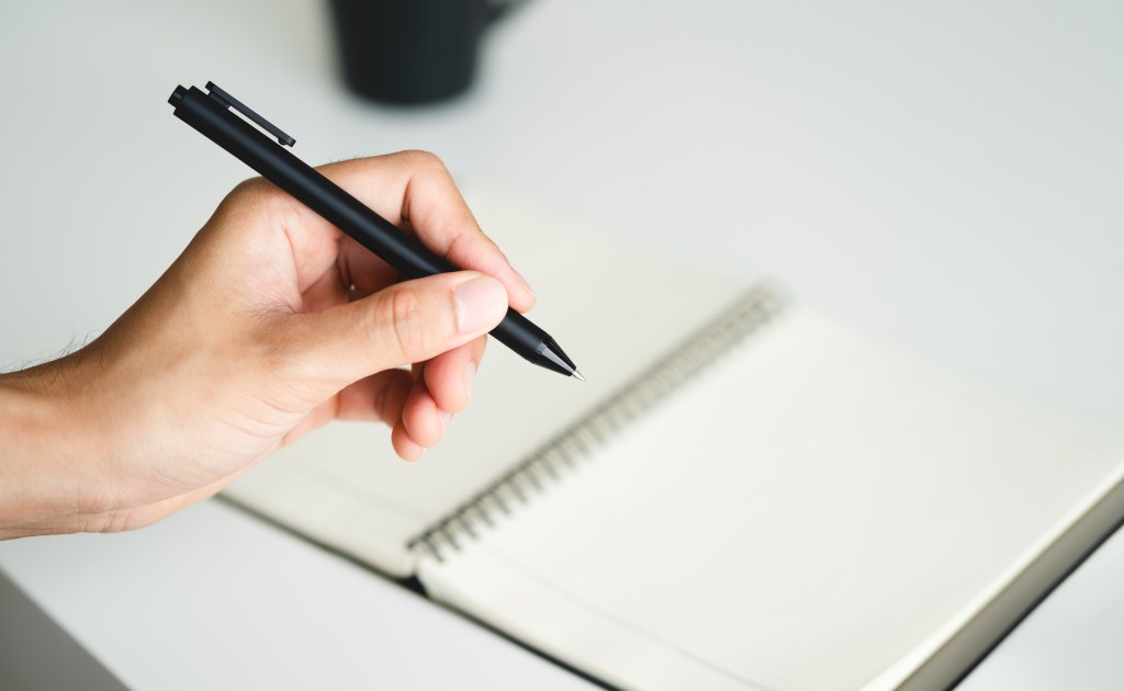 Left-handed man holding a pen and writing in a notebook on a table