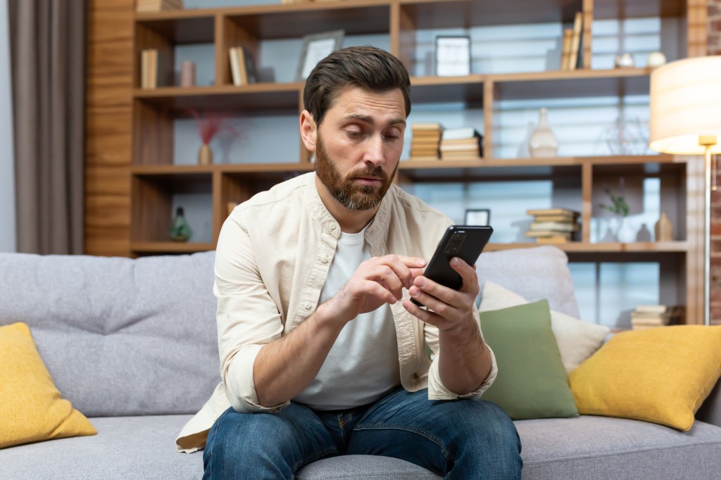 Man resembling Erik Gudbranson sitting bored on a couch at home, scrolling through dating apps on his phone