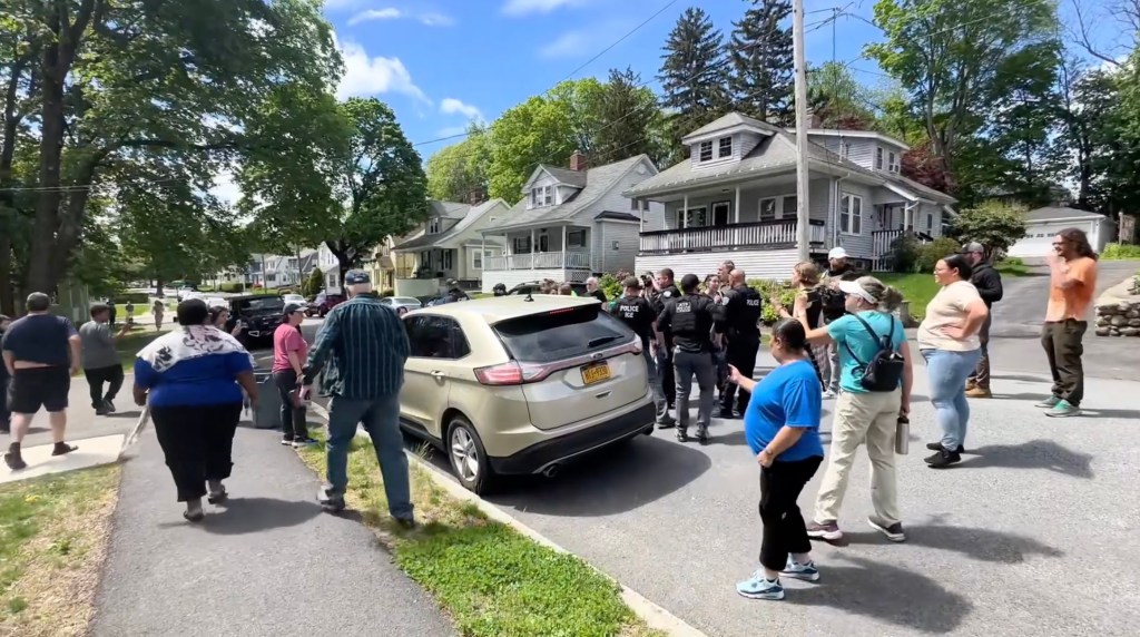 Crowd of approximately 25 people in Massachusetts protesting an ICE arrest, with some individuals placing hands on an SUV with a detained woman inside, amidst a tense confrontation with uniformed ICE agents.