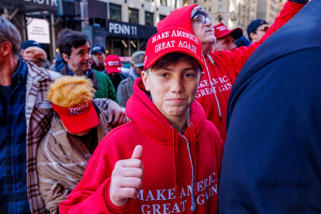 A young person wearing a "Make America Great Again" hat and sweatshirt gives a thumbs-up at a political rally.