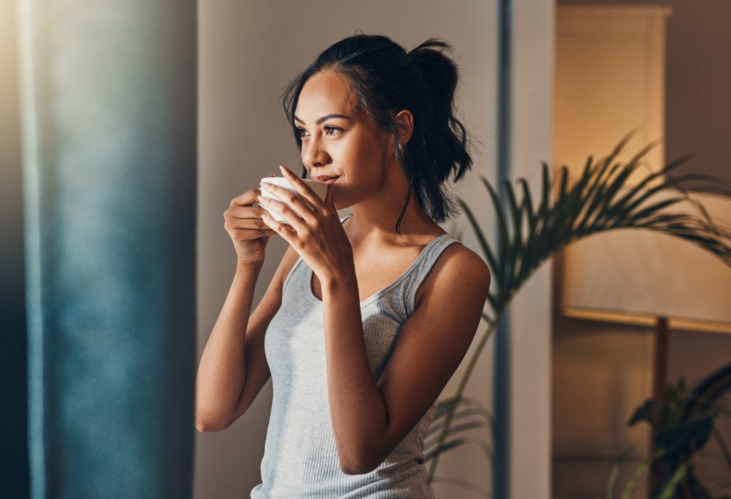 A new study out of Harvard found that caffeinated coffee consumption in midlife can lead to healthy aging. Woman drinking coffee by a window.