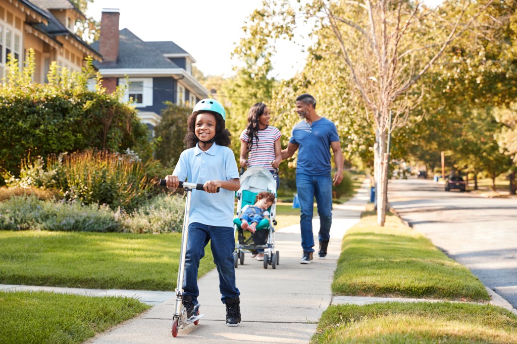 Family walking down a sidewalk; a child rides a scooter while parents push a stroller with a baby.
